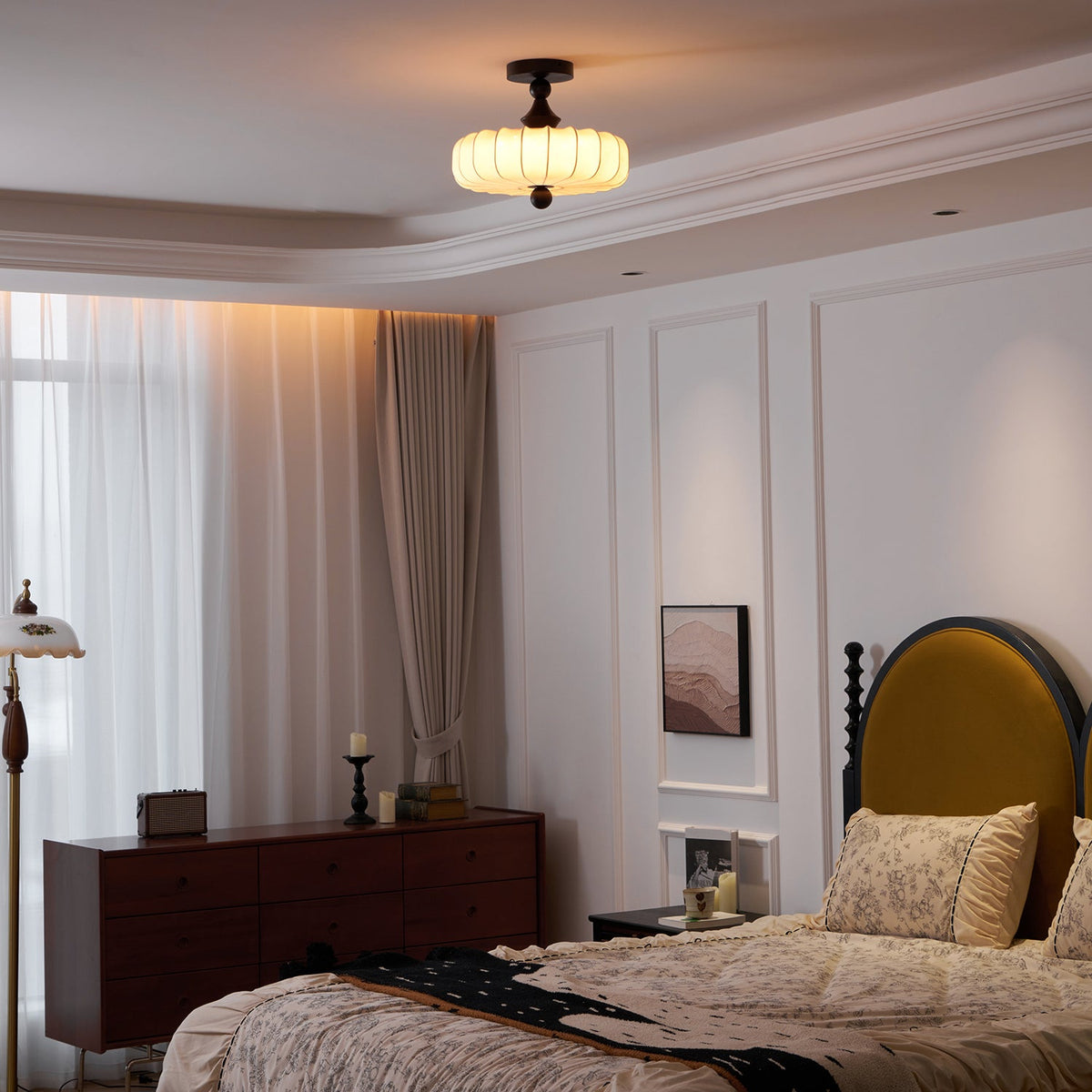 A Clérachic ceiling light with a ribbed, glowing shade hangs above a bed with a mustard headboard, beside a wooden chest of drawers.