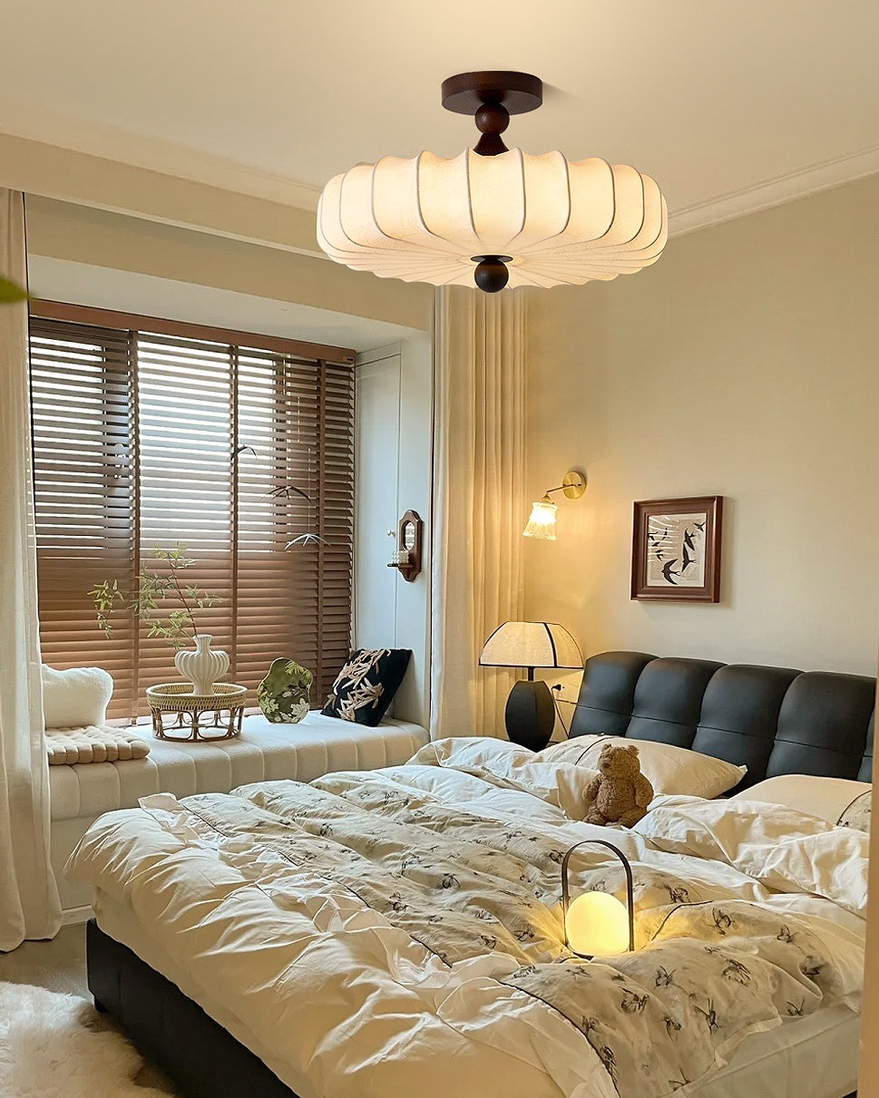 A Clérachic ceiling light with a pleated cream shade hangs above a bed with a dark headboard and a bright window with wooden blinds.