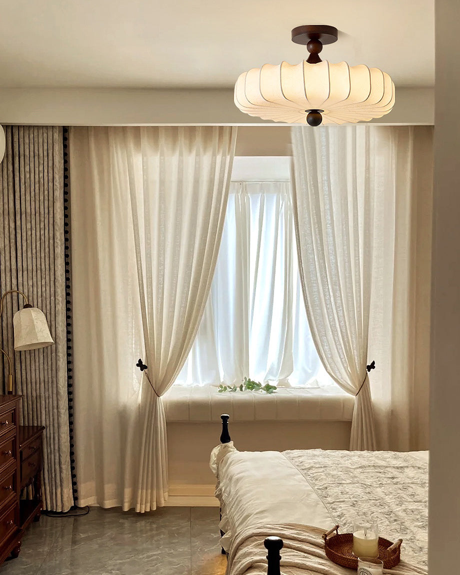A Clérachic ceiling light with a soft pleated shade hangs by sheer curtains, above a bed with a floral cover and a small side table.