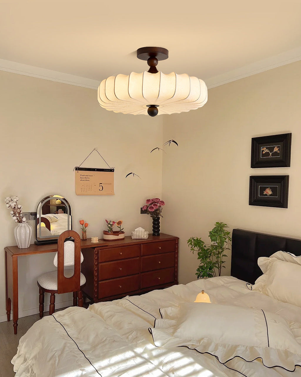 A Clérachic ceiling light glows above a bedroom with a wooden dresser, a small dressing mirror and a bed made up in white linen.