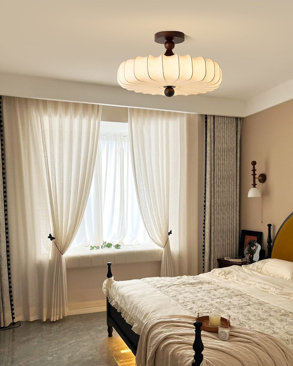 Beside sheer cream curtains, a Clérachic ceiling light with a pleated shade hangs above a bed with a dark frame and a knitted throw.