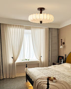 Beside sheer cream curtains, a Clérachic ceiling light with a pleated shade hangs above a bed with a dark frame and a knitted throw.