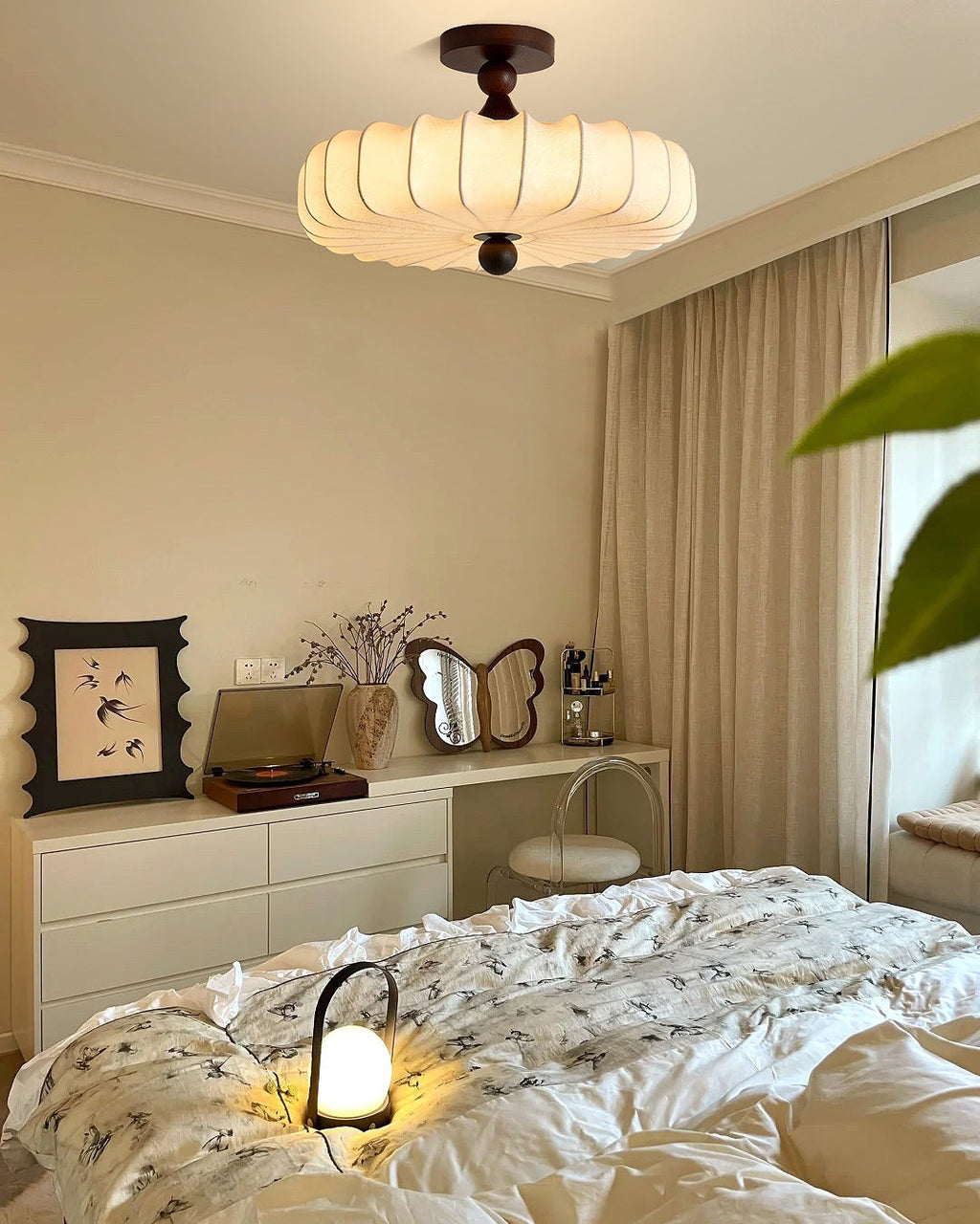 A lit Clérachic ceiling light hangs above a white dresser and a bed with a printed duvet, with a small glowing lamp on the cover.