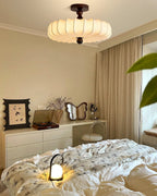 A lit Clérachic ceiling light hangs above a white dresser and a bed with a printed duvet, with a small glowing lamp on the cover.