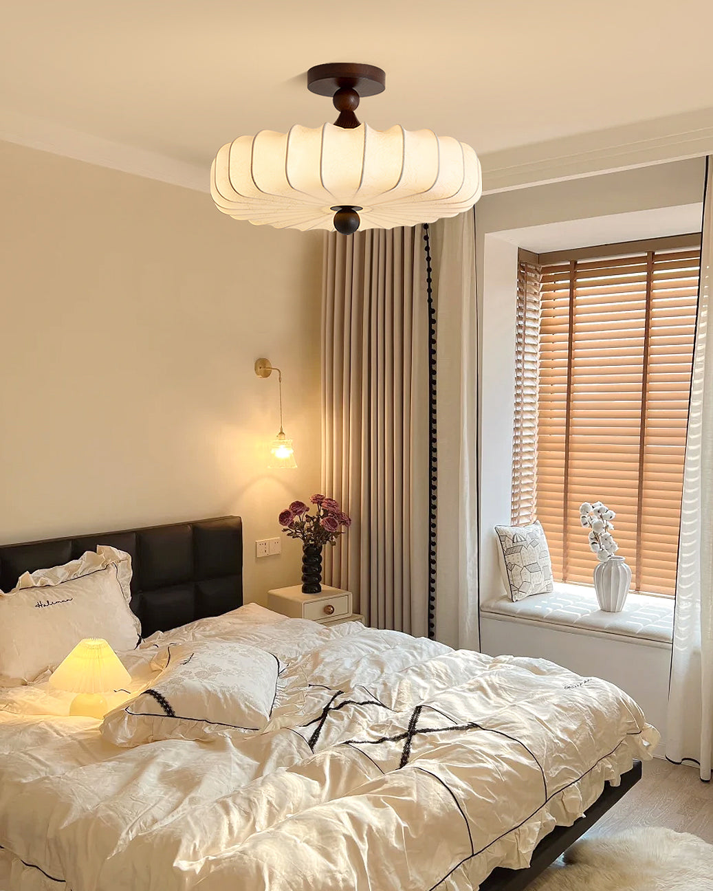 A Clérachic ceiling light hangs near a window with wooden blinds, above a bed with a black padded headboard and a small wall lamp.