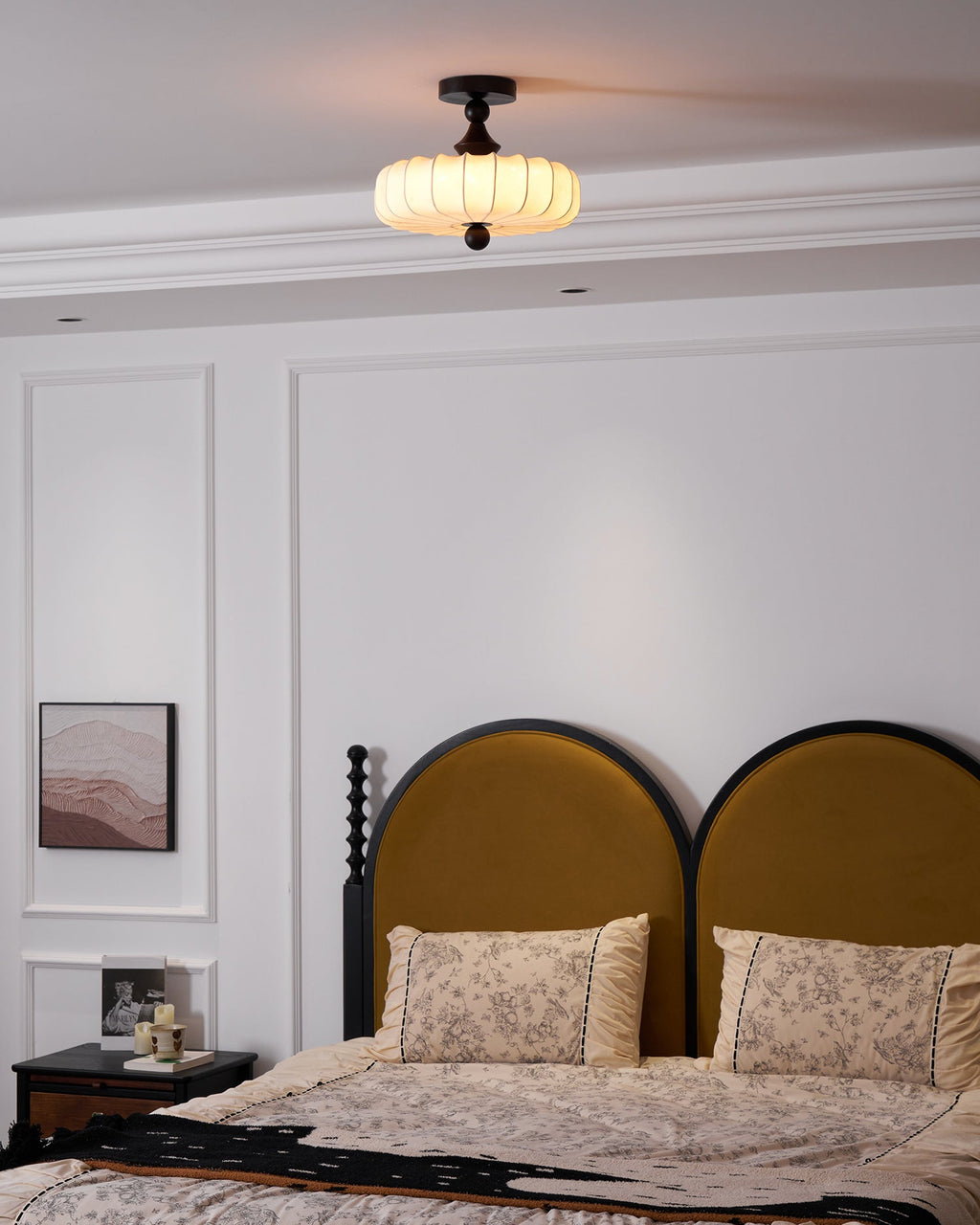 Seen from the foot of the room, a small Clérachic ceiling light hangs above a bed with a mustard arched headboard and floral pillows.
