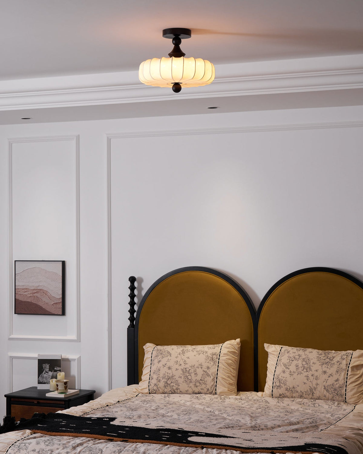 Seen from the foot of the room, a small Clérachic ceiling light hangs above a bed with a mustard arched headboard and floral pillows.