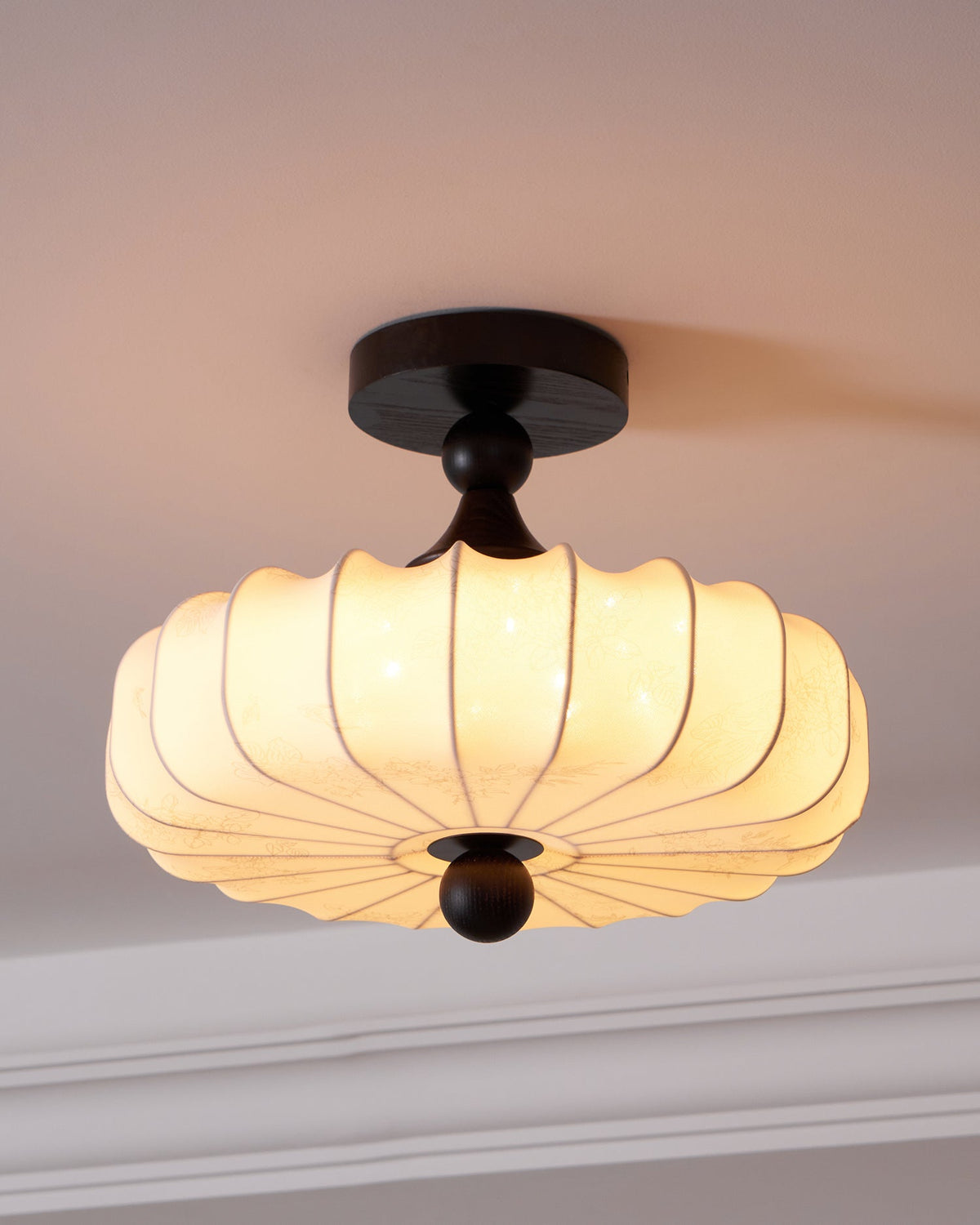Close view of a Clérachic ceiling light with a lit, pleated beige shade, a dark walnut-colour canopy and a round wooden finial.