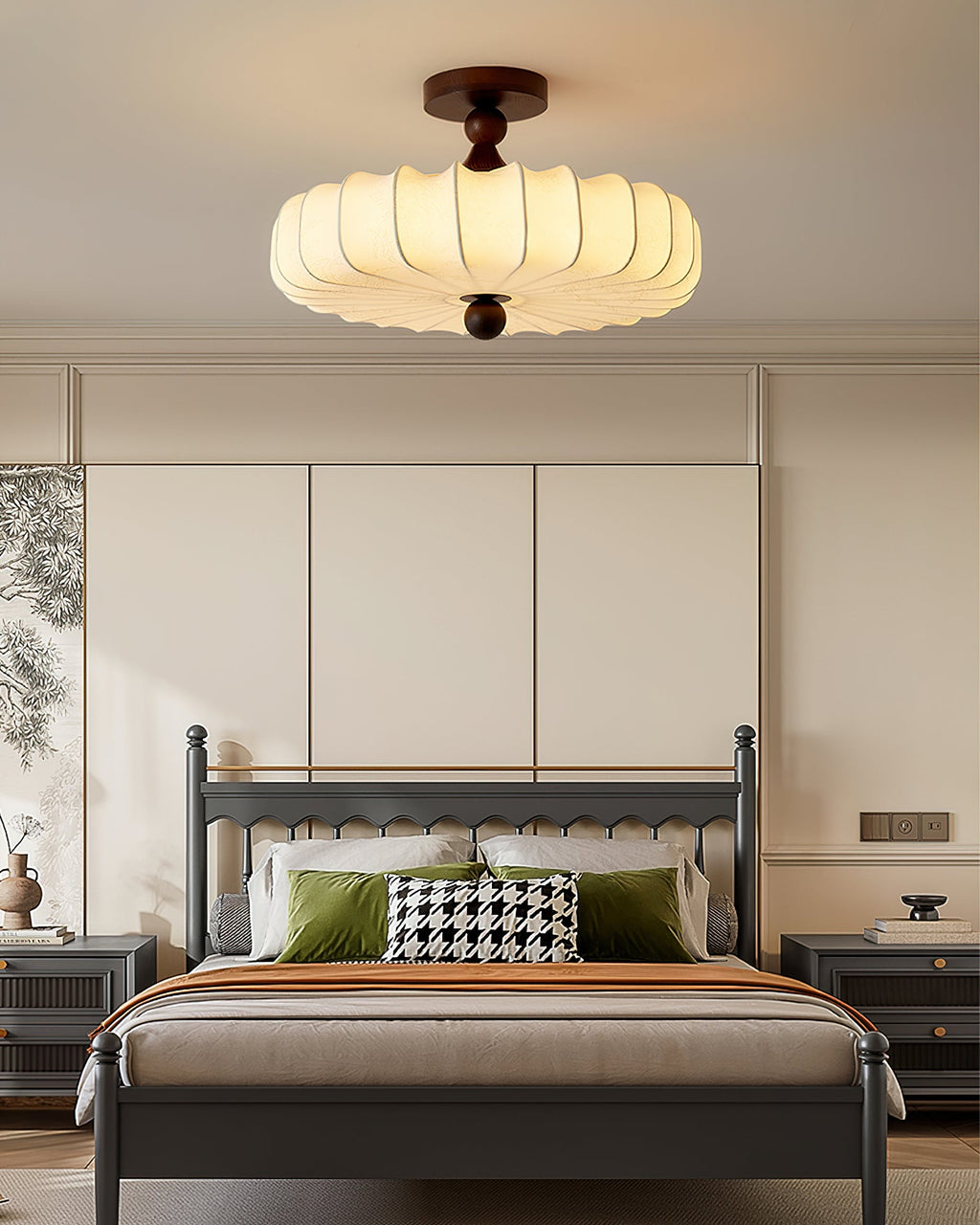 A Clérachic ceiling light with a glowing pleated shade hangs above a grey-framed bed, with green cushions and a houndstooth pillow below.