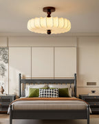 A Clérachic ceiling light with a glowing pleated shade hangs above a grey-framed bed, with green cushions and a houndstooth pillow below.