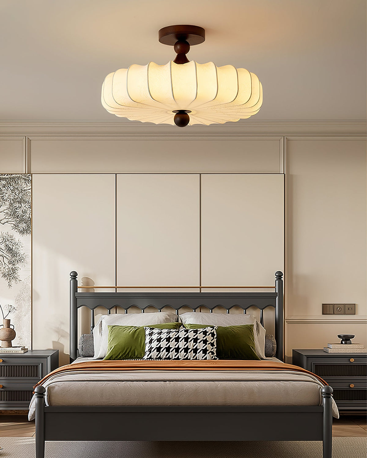 A Clérachic ceiling light with a glowing pleated shade hangs above a grey-framed bed, with green cushions and a houndstooth pillow below.