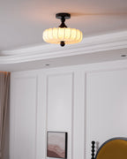 A lit Clérachic ceiling light with a pleated shade hangs near a panelled wall with a framed print and the edge of a mustard headboard.