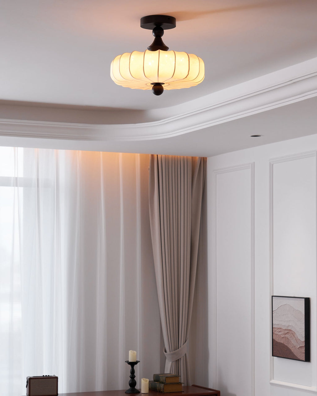 A small Clérachic ceiling light glows warm in a room with a stepped ceiling, long sheer curtains and a framed print on the wall.
