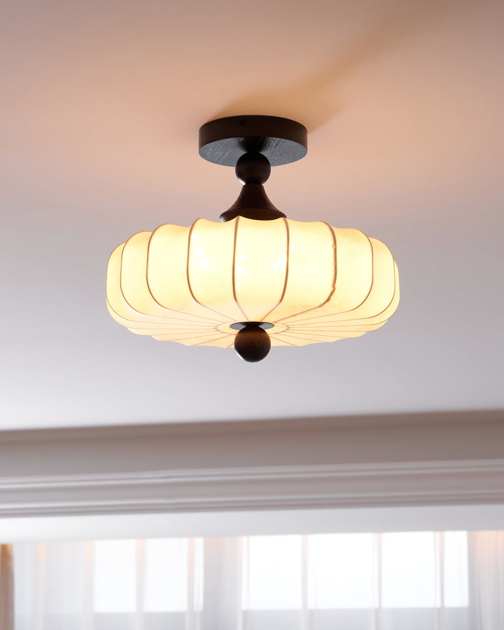 A Clérachic ceiling light with a pleated, glowing shade hangs beneath a white moulded ceiling, with bright windows in the background.