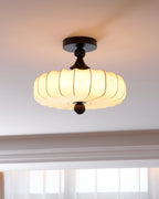 A Clérachic ceiling light with a pleated, glowing shade hangs beneath a white moulded ceiling, with bright windows in the background.