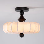 Side view of a Clérachic ceiling light, with a dark round canopy, a turned wooden stem and a wide pleated fabric drum, softly lit.