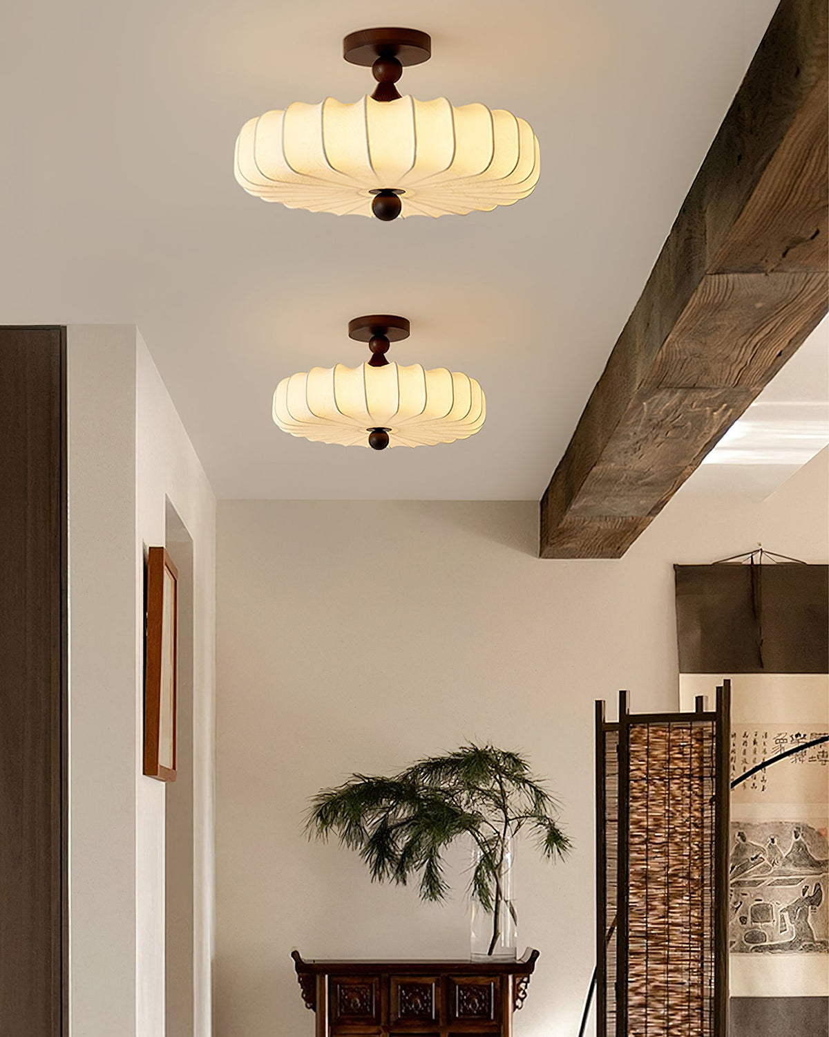 Two Clérachic ceiling lights, lit warm, hang in a hallway beside a rough timber beam, with a carved console and a woven screen beyond.