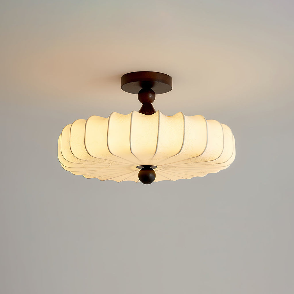 A lit Clérachic ceiling light with a wide pleated shade hangs on a pale grey backdrop, a small round finial below.