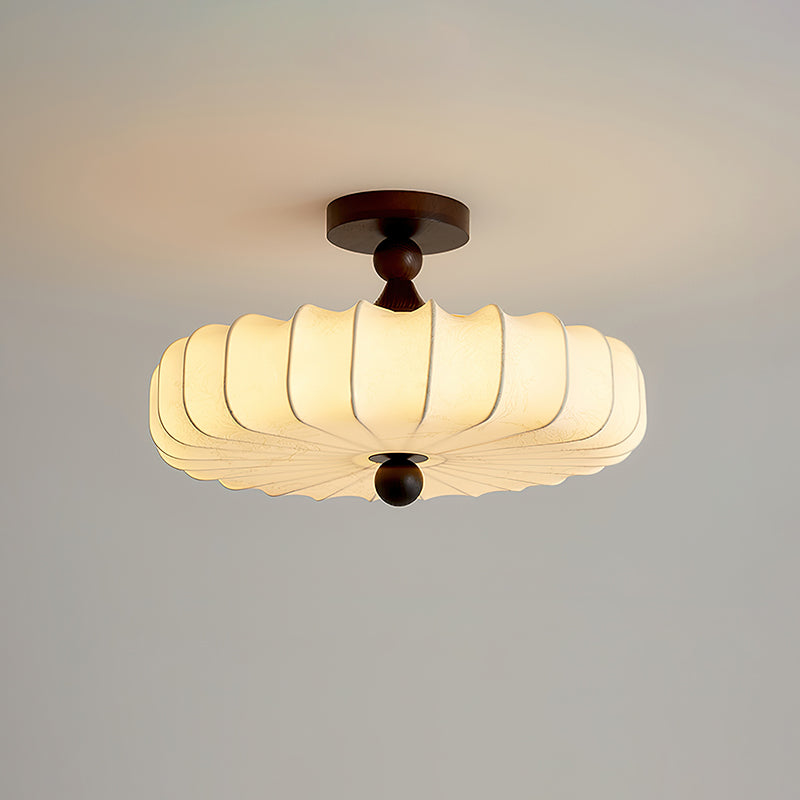 A broad Clérachic ceiling light with a lit, ribbed fabric shade and a round finial hangs against a grey and peach backdrop.