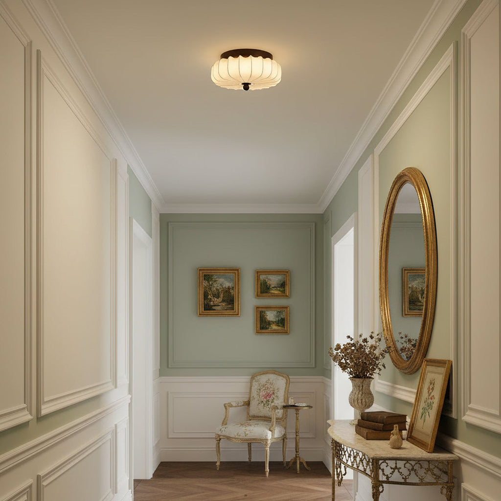 Above a panelled hallway with a gilt mirror and a small cream armchair at the end, this pleated Clérachic ceiling light glows.