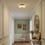 Above a panelled hallway with a gilt mirror and a small cream armchair at the end, this pleated Clérachic ceiling light glows.