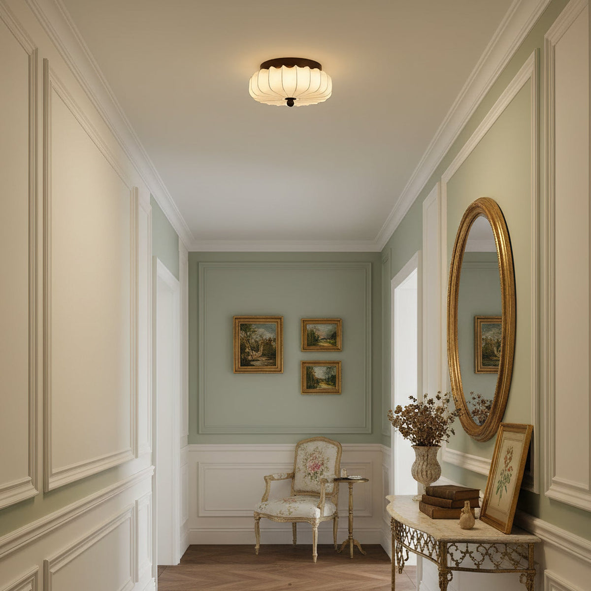 Above a panelled hallway with a gilt mirror and a small cream armchair at the end, this pleated Clérachic ceiling light glows.