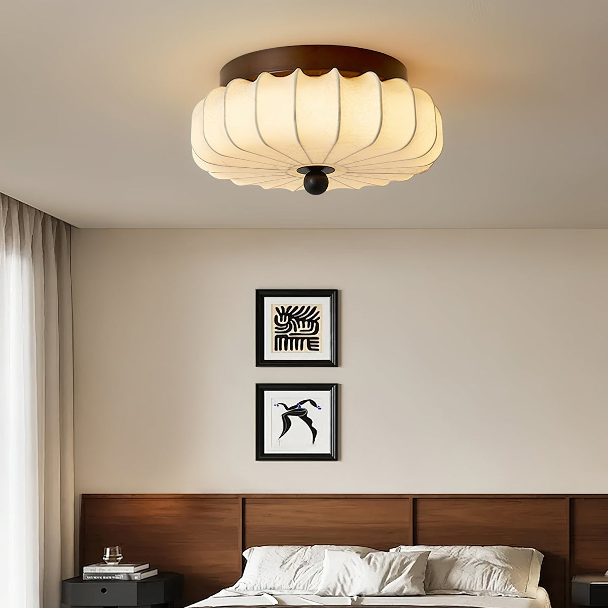 Above a bed with a walnut headboard and two framed prints, a large pleated fabric Clérachic ceiling light glows warmly.