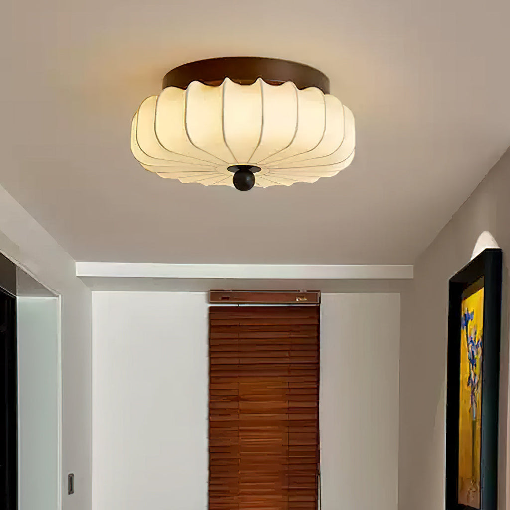 Seen close up above a hallway with a timber blind and a framed painting, this pleated fabric Clérachic ceiling light is lit.