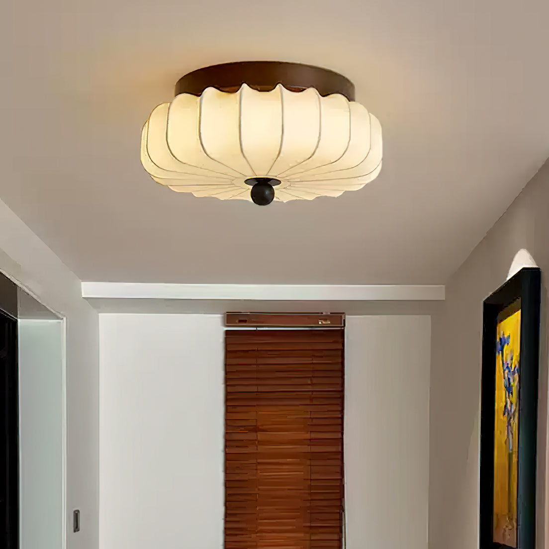 Seen close up above a hallway with a timber blind and a framed painting, this pleated fabric Clérachic ceiling light is lit.