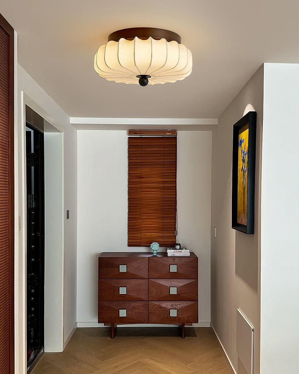 In a narrow hallway with a walnut chest of drawers and a timber blind beyond, this pleated Clérachic ceiling light is lit.