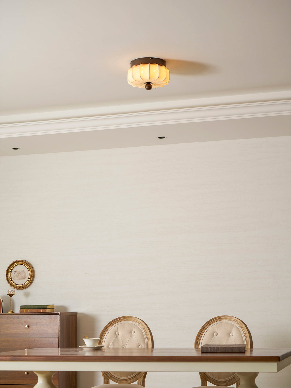 A small Clérachic ceiling light lit warm above a dining room with a long table, carved-back chairs and a cornice.