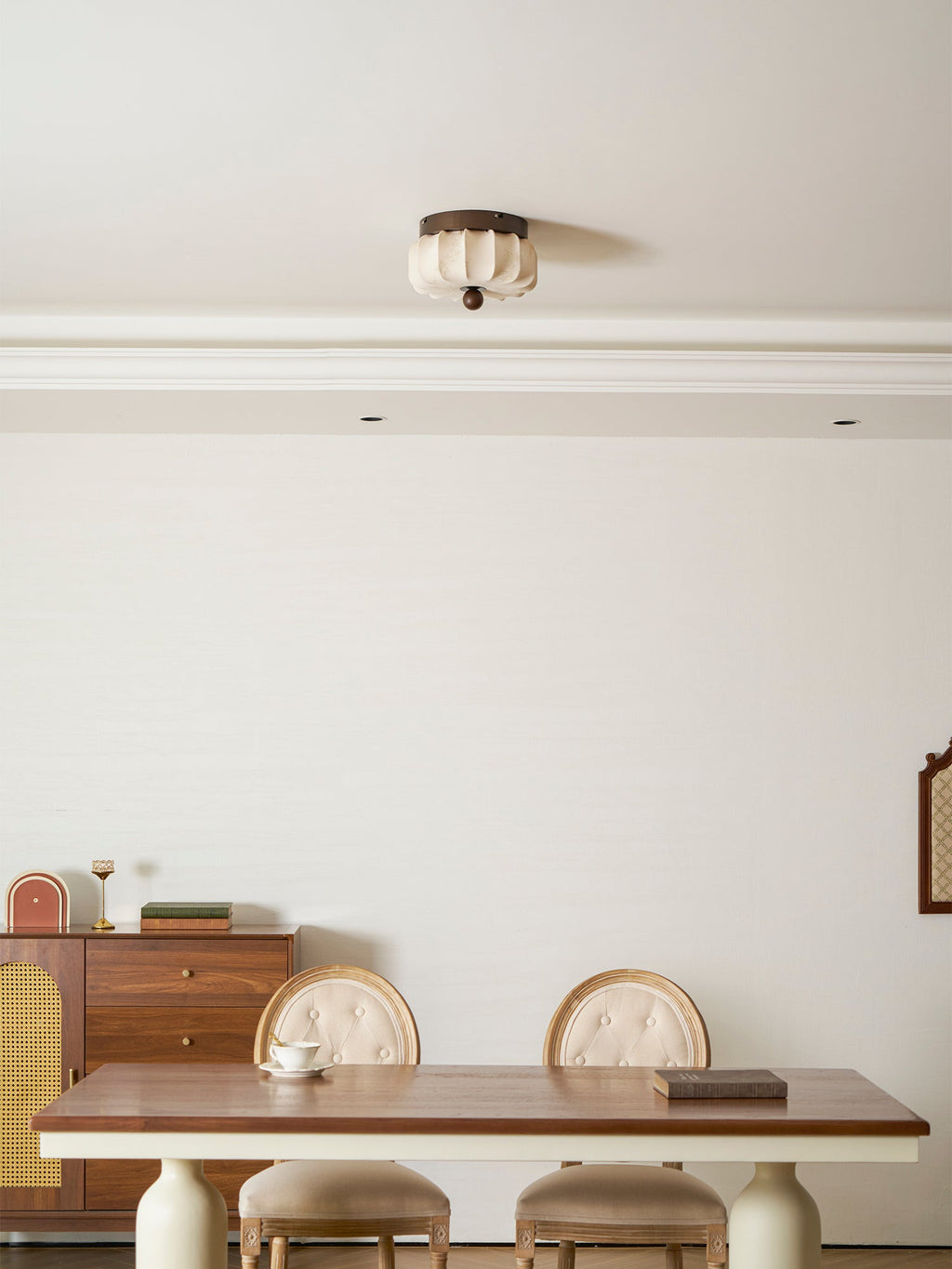 A small pleated Clérachic ceiling light, unlit, high above a dining table, with a rattan-fronted sideboard to the left.
