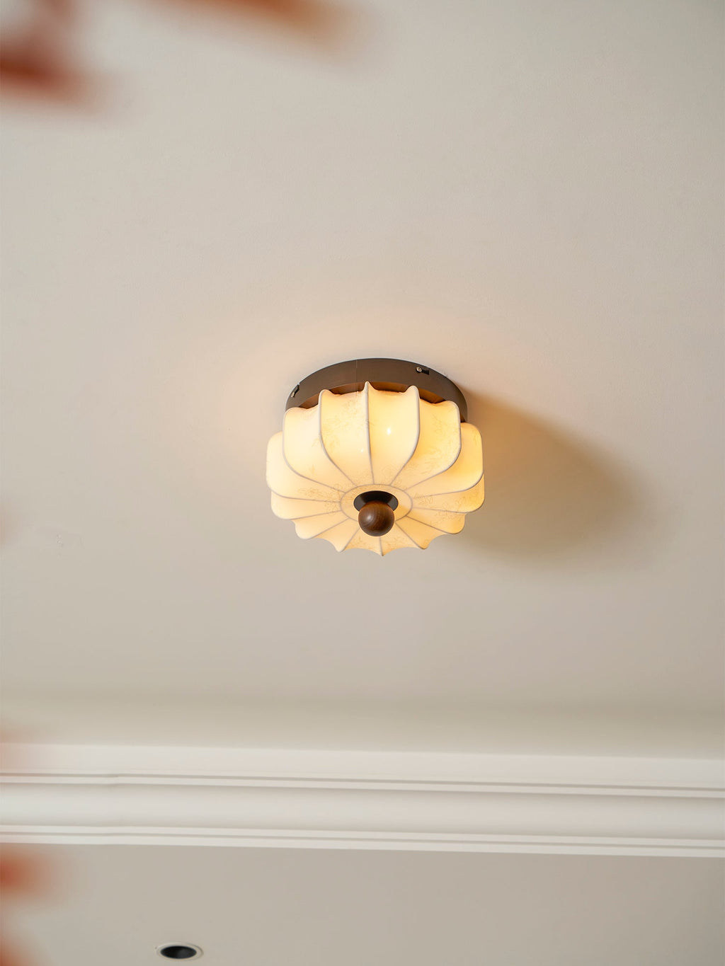 A pleated fabric Clérachic ceiling light lit warm, seen from below, with a cornice and blurred red leaves in the corner.