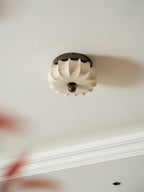 A pleated fabric Clérachic ceiling light seen from below at an angle, unlit, with blurred red leaves at the lower left.