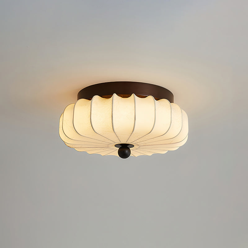 A pleated fabric Clérachic ceiling light lit, with a warm glow around its dark walnut frame, against a grey ceiling.