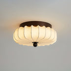 A pleated fabric Clérachic ceiling light lit, with a dark frame and a wooden ball below, against a grey ceiling.