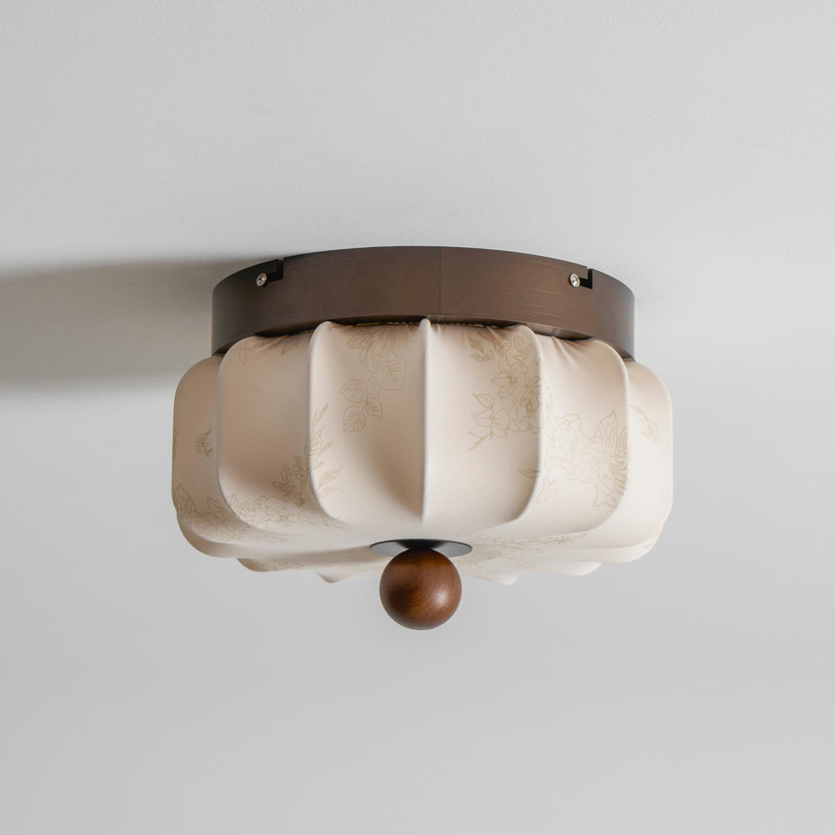 A pleated fabric Clérachic ceiling light, unlit, with a faint floral print on the fabric and a walnut ball below.