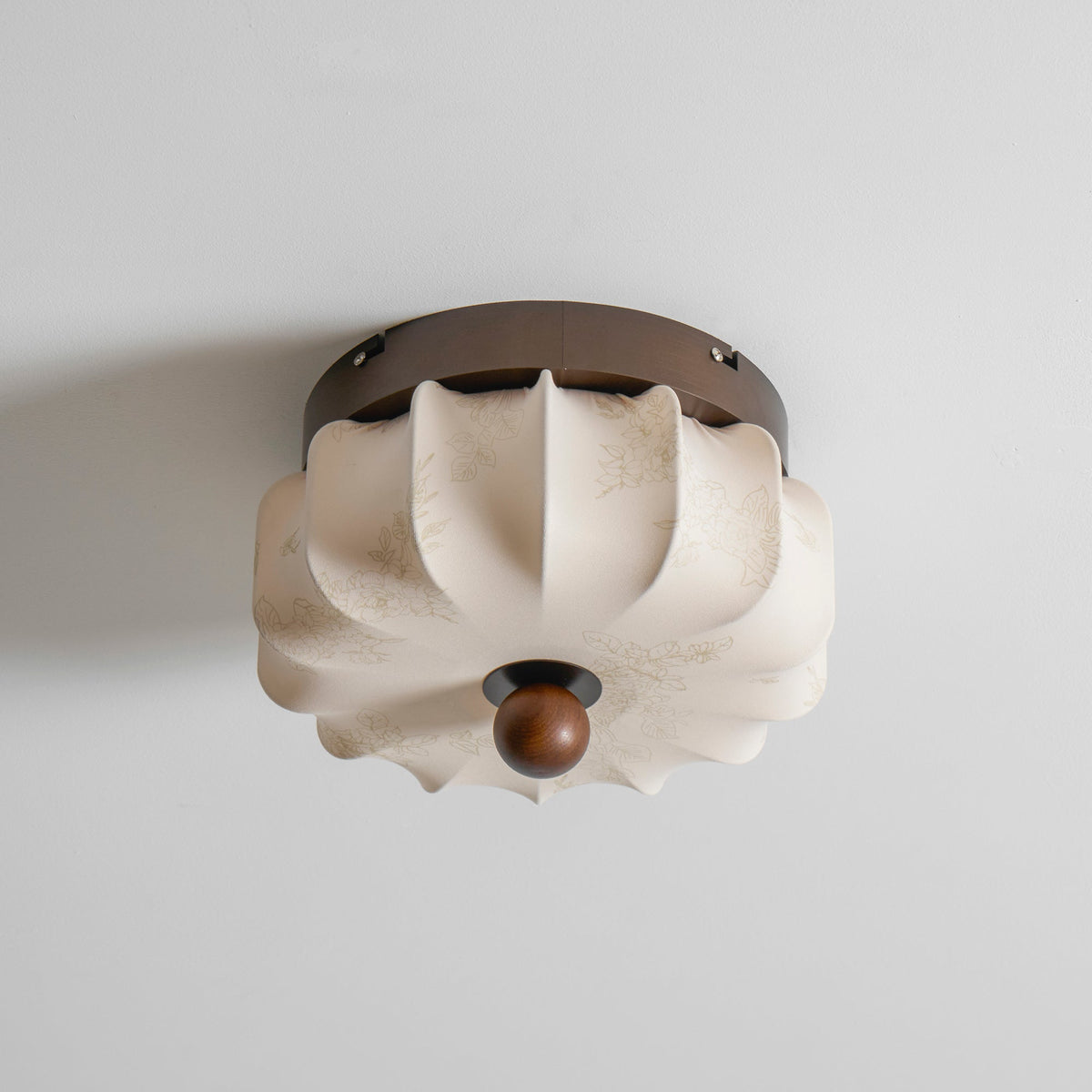 A pleated fabric Clérachic ceiling light, unlit, with a folded shade and a faint floral print, on a grey ceiling.