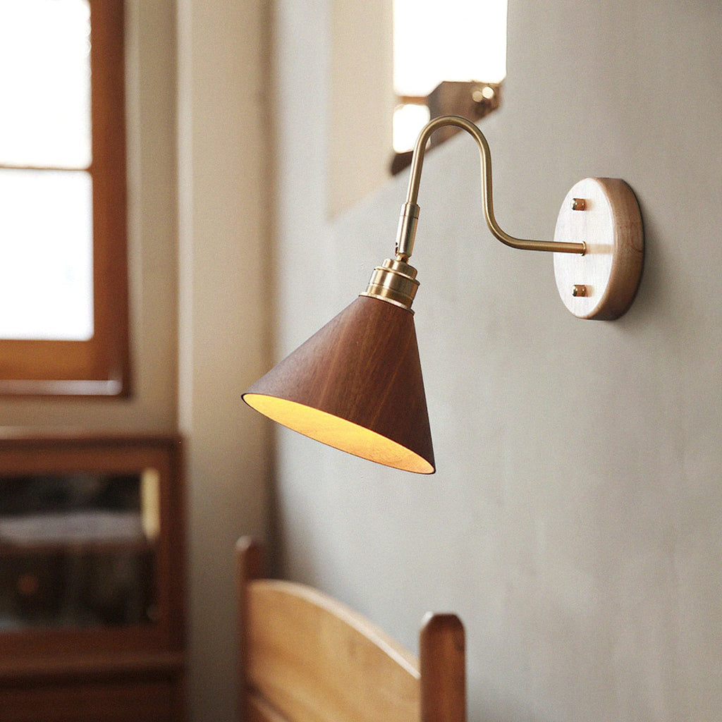 A Clérachic wall light with a walnut-colour cone shade glowing warmly against a grey plaster wall beside a sunlit window.