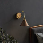 Small Clérachic wall light with a walnut-colour cone shade and brass arm on a dark charcoal wall above a leather headboard.