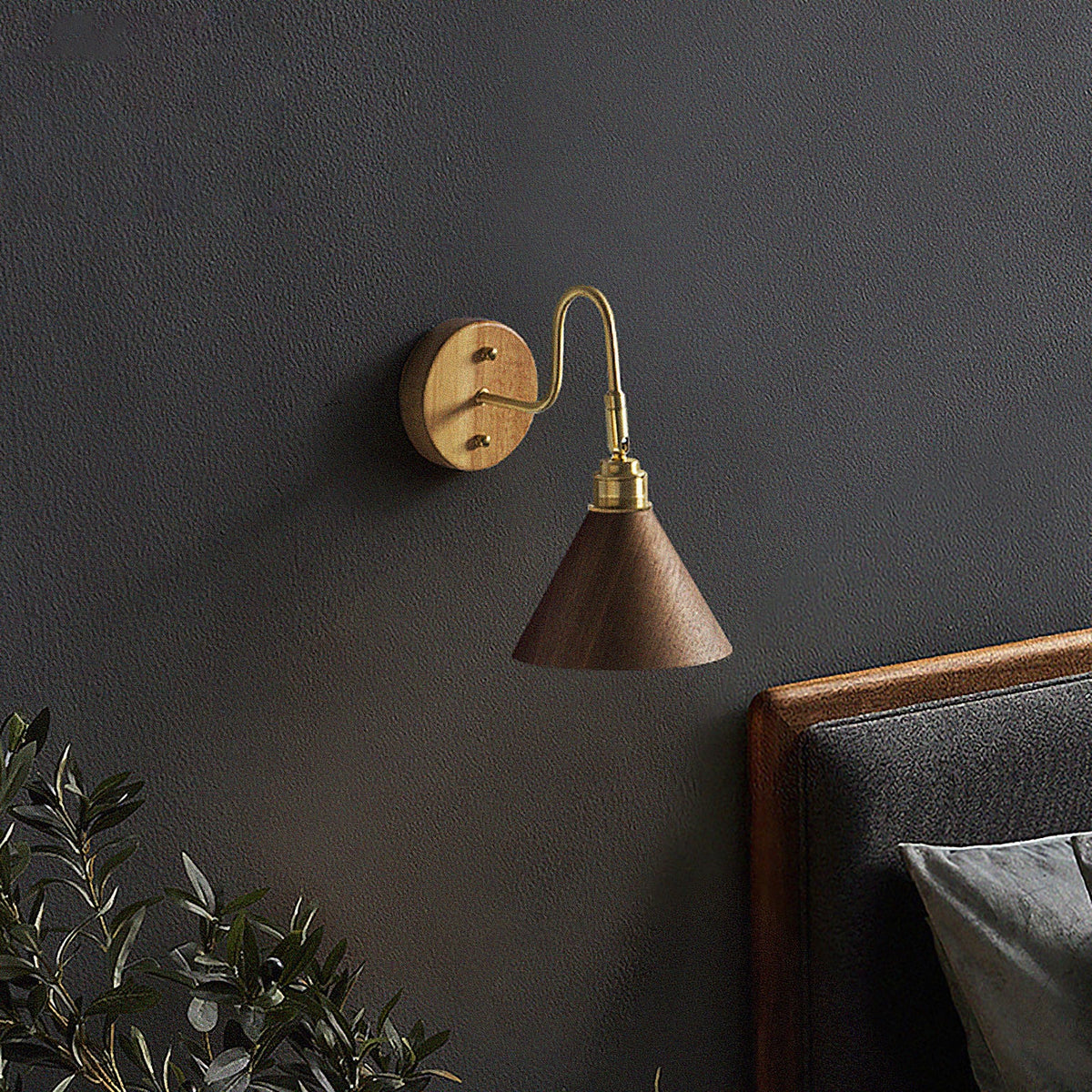 Small Clérachic wall light with a walnut-colour cone shade and brass arm on a dark charcoal wall above a leather headboard.