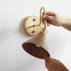 A hand holds the brass lamp holder while another lifts the walnut-colour cone shade of this Clérachic wall light.