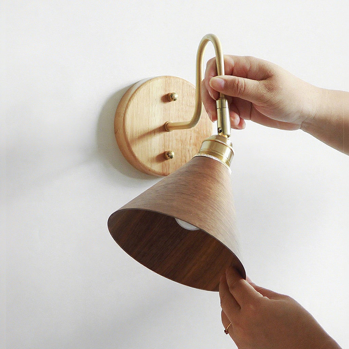 A hand holds the brass lamp holder while another lifts the walnut-colour cone shade of this Clérachic wall light.