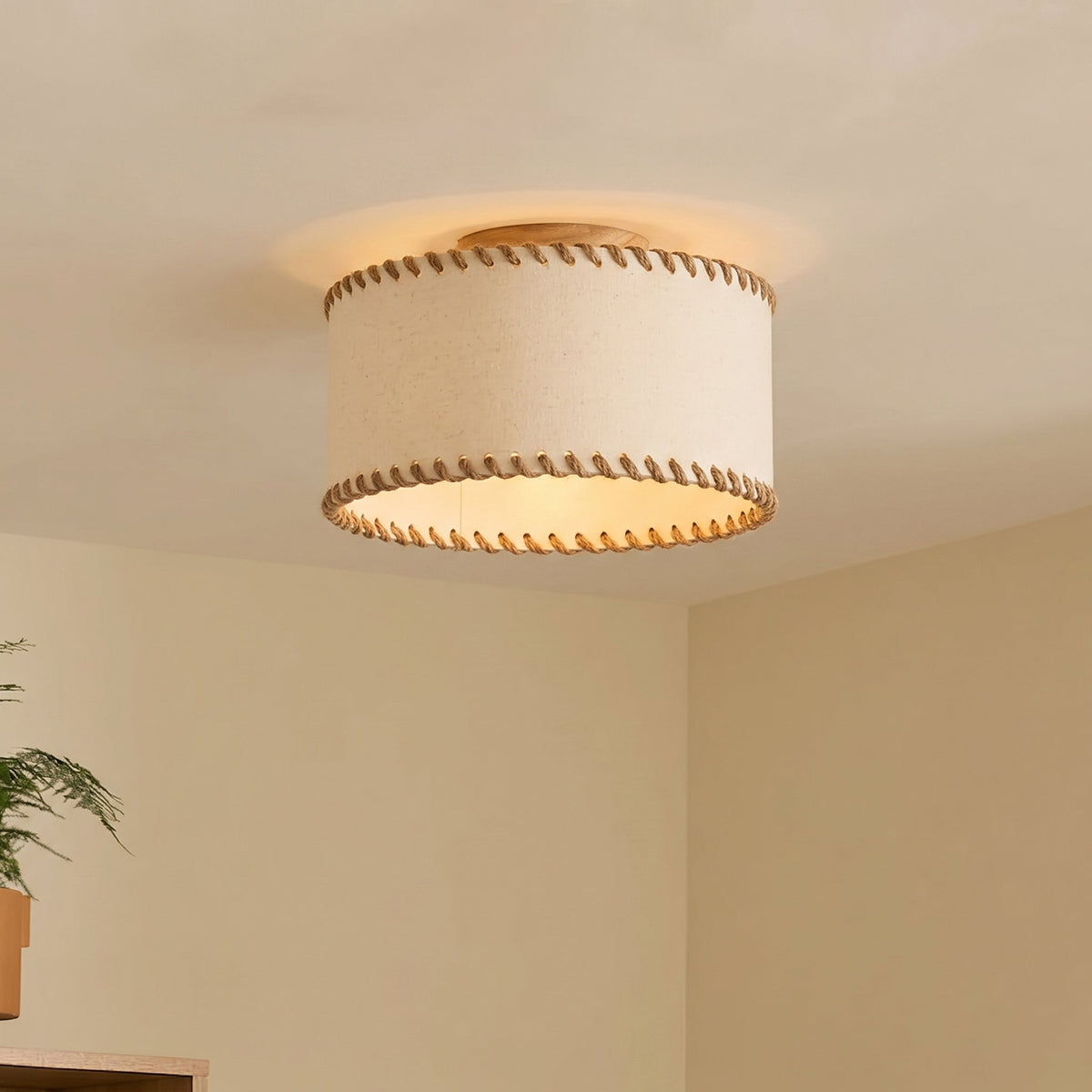Lit drum shade with rope-stitched edges on a ceiling in the corner of a cream room, a Clérachic flush mount casting a warm halo around its wood canopy.