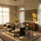 Clérachic wavy ribbon chandelier glowing over a long dark wood dining table with cane-backed chairs, sheer curtains and a vase of flowers.