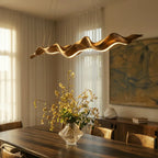 Clérachic wavy wood-finish chandelier close up above a dining table, its warm LED line glowing beside a vase of yellow blossom.