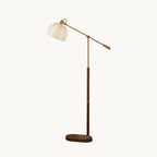 Walnut colour Clérachic floor lamp on a plain white backdrop, with a pleated beige dome shade on a long brass arm and an oval wooden base.