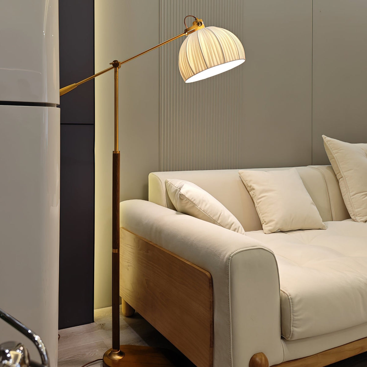 This Clérachic floor lamp leans over a cream sofa with plump cushions, its beige shade lit against a dark panel and a fridge edge.