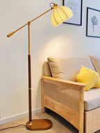 Walnut colour Clérachic floor lamp behind a rattan-sided wooden sofa with a yellow cushion, the shade lit and angled over the seat.