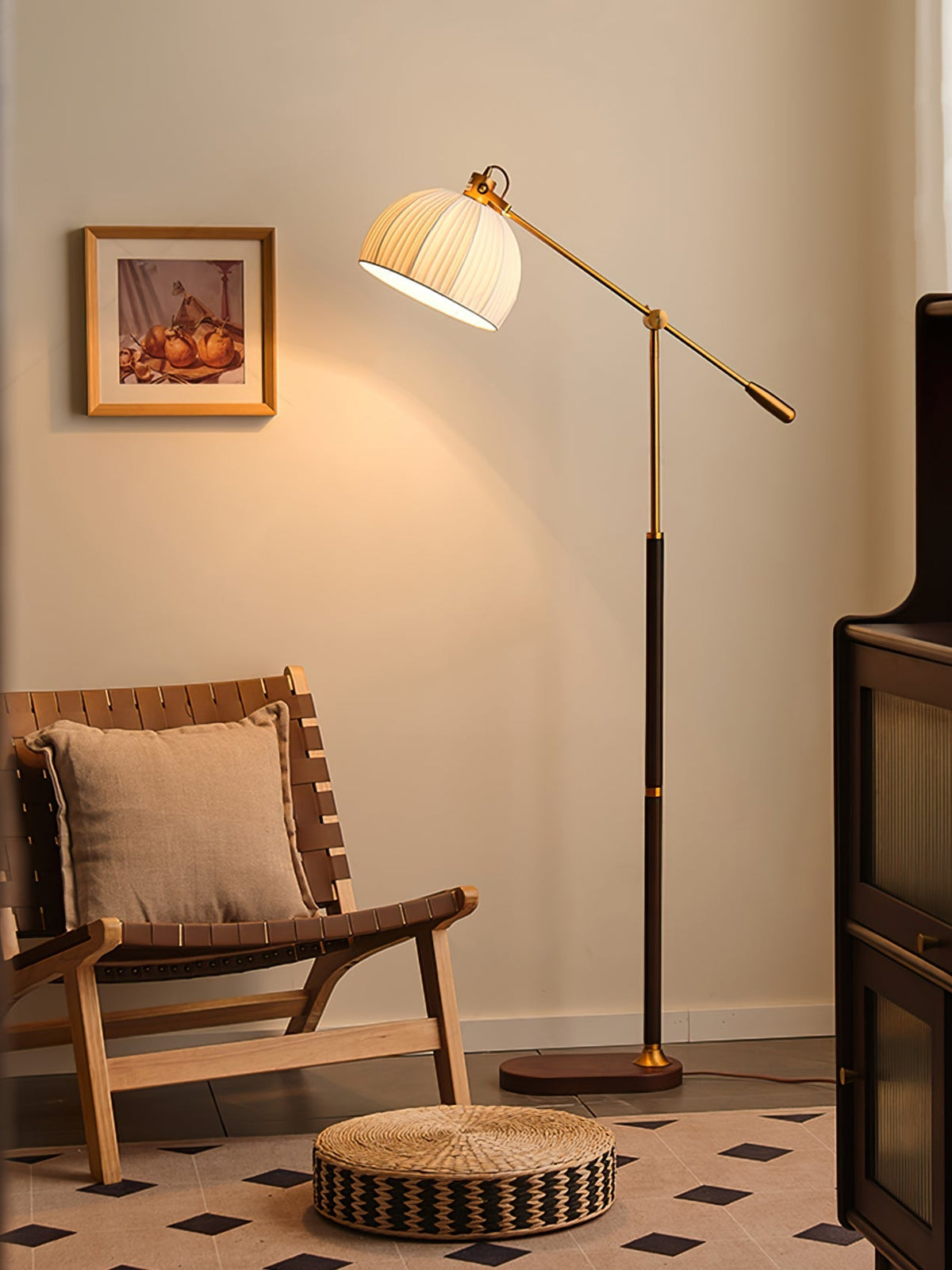 Clérachic floor lamp lit in a warm room, beside a woven wooden chair, a round straw pouffe and a black and white tiled floor.
